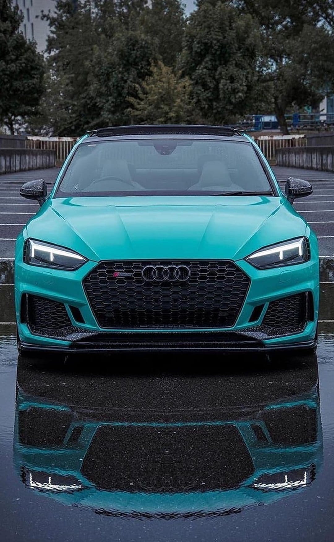 Turquoise Audi RS5 sports car on reflective wet asphalt with an aggressive front grille and glowing LED lights.