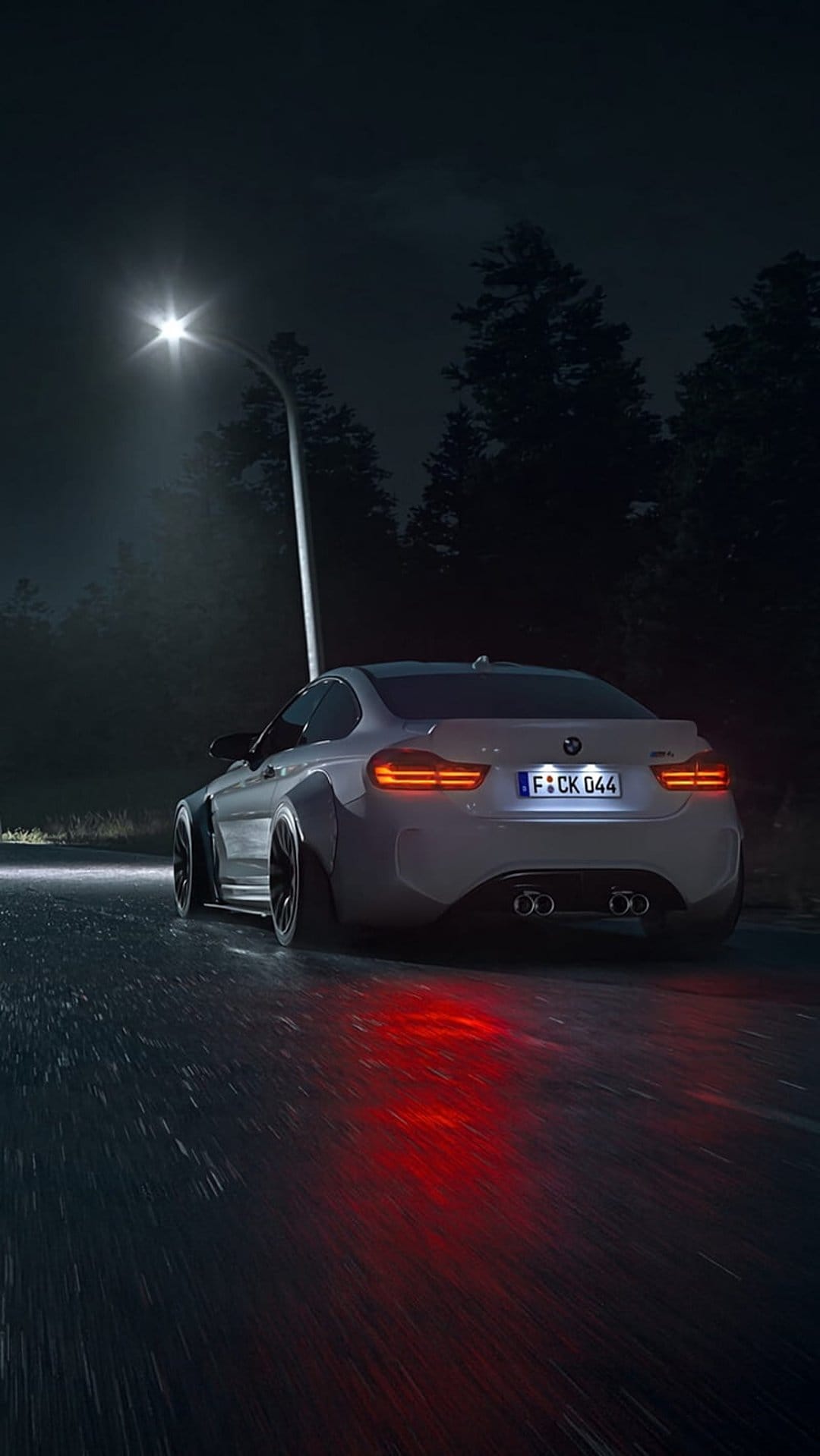 White BMW M4 coupe with widebody kit driving on wet asphalt at night under red taillight reflections.