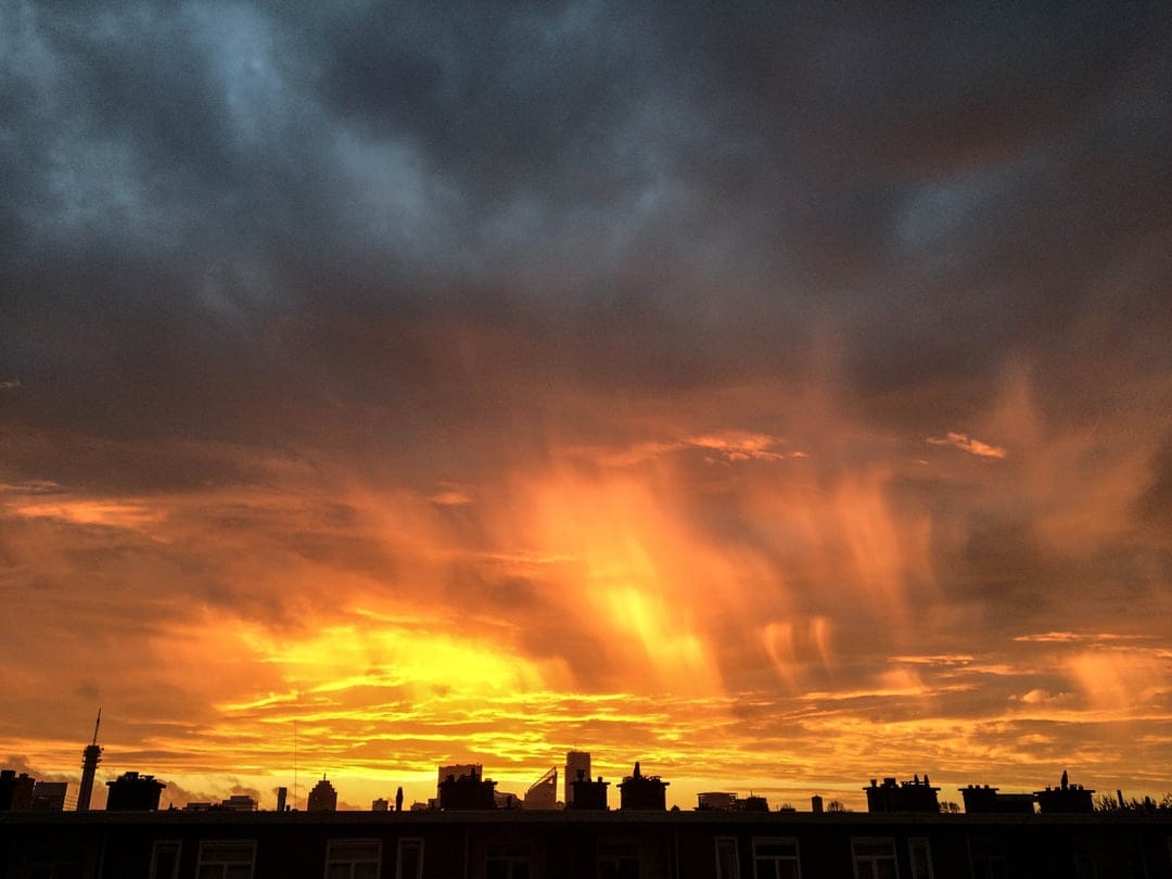 Sunbeams burst through dark orange and red clouds above the black silhouette of a distant city skyline at dusk.