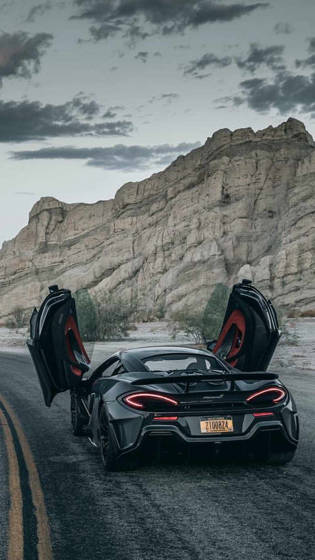 McLaren supercar with open dihedral doors parked on a desert road against rugged rocks and a cloudy sky.