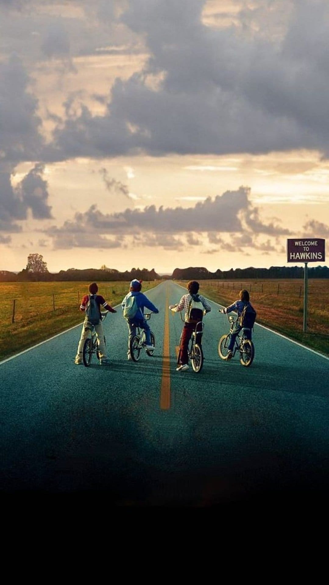 Four young friends ride bicycles on a road toward a Hawkins sign under a dark, dramatic, stormy sky.