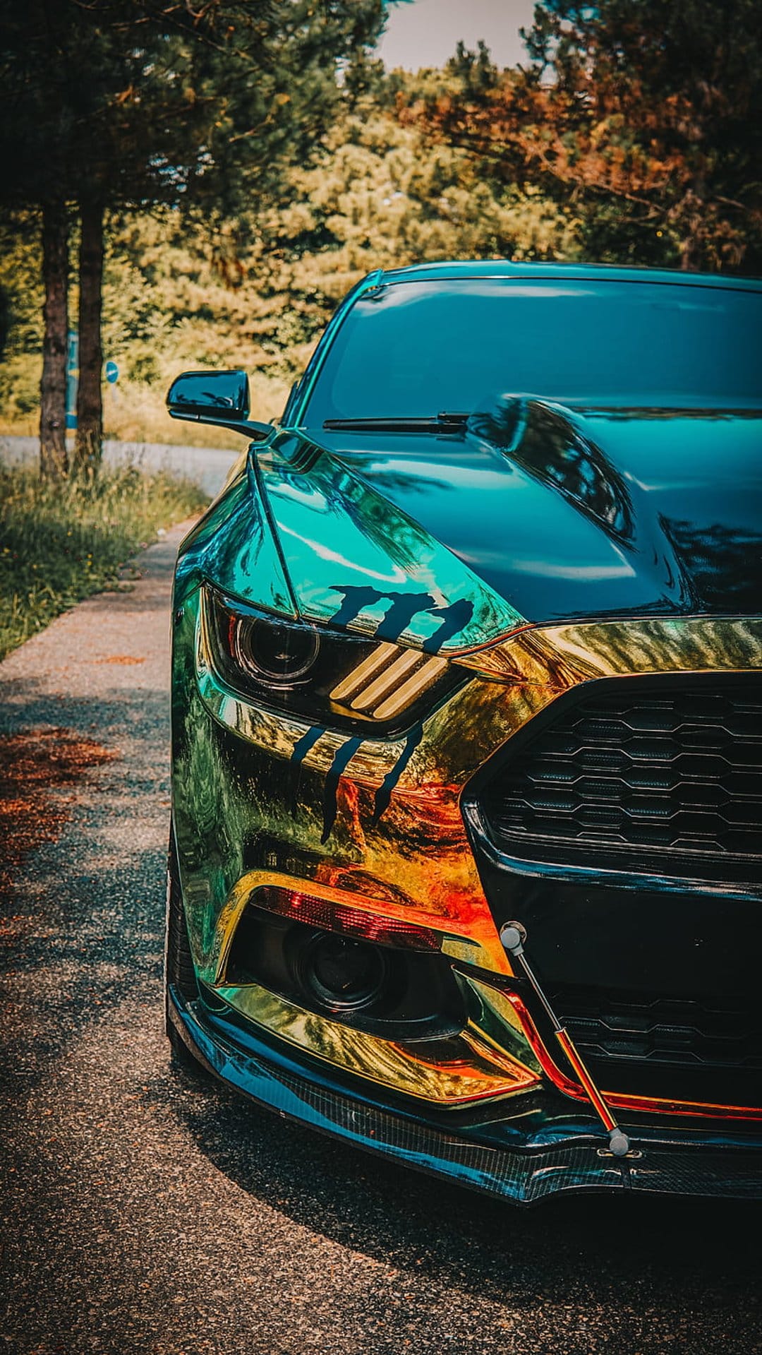 Ford Mustang with an iridescent chroma wrap and red tow straps parked on an asphalt path near green trees.
