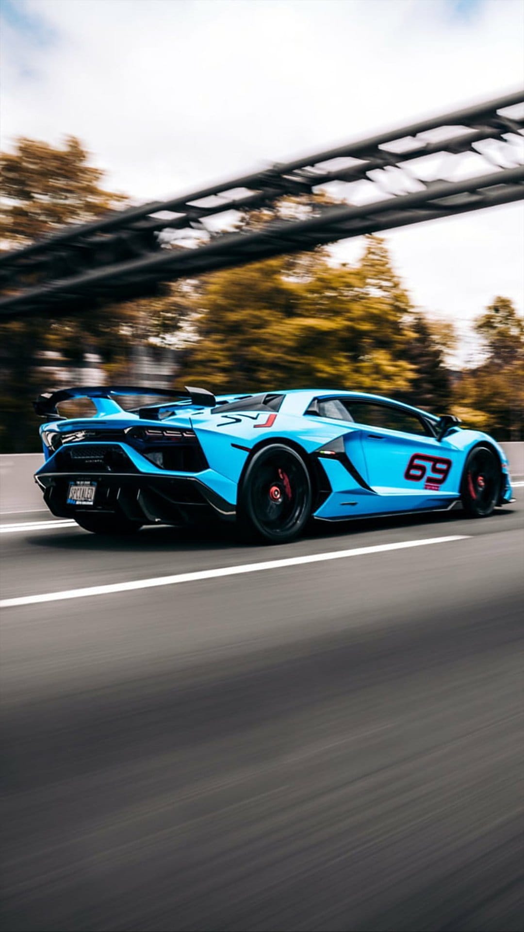 Blue Lamborghini Aventador SVJ with number 69 racing on a highway past autumn trees and a concrete overpass.