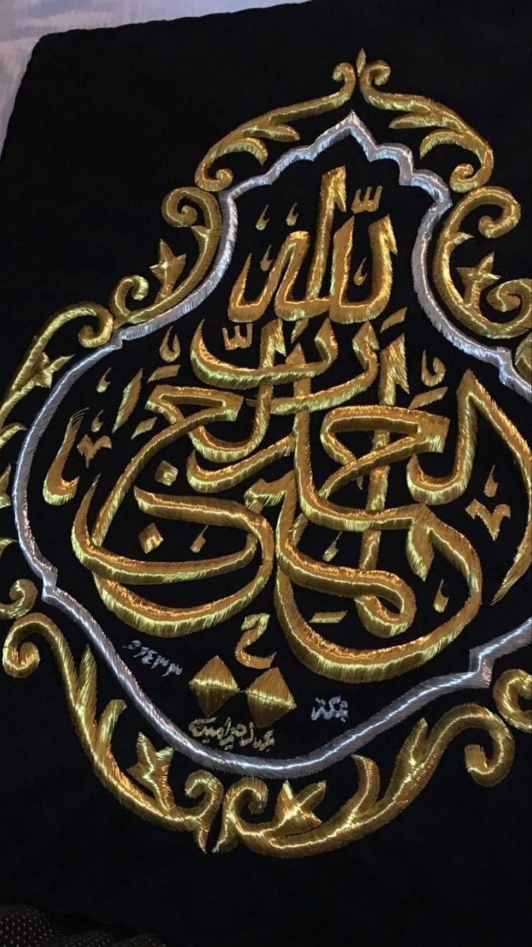 Gold and silver embroidered Arabic calligraphy on a textured black fabric background with ornate floral details.