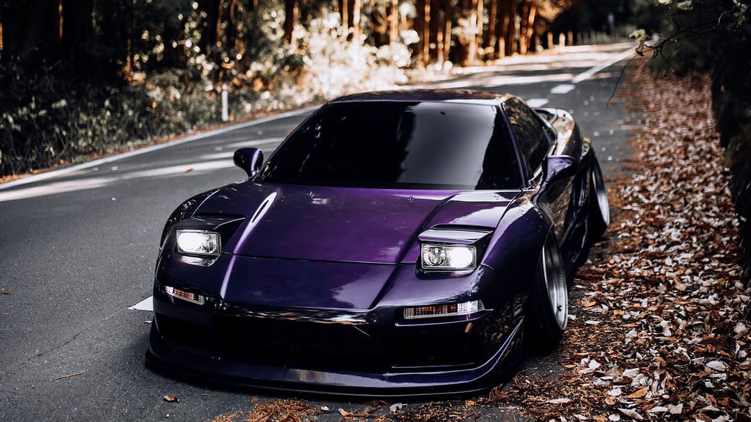 Purple widebody NSX sports car with illuminated headlights parked on a road with autumn leaves and forest bokeh.