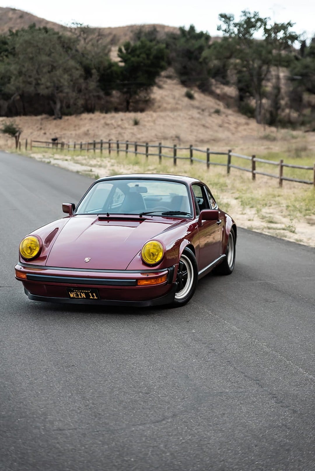 Maroon Porsche 911 with yellow headlights sits on a paved road beside a wooden fence and dry grassy hillsides.