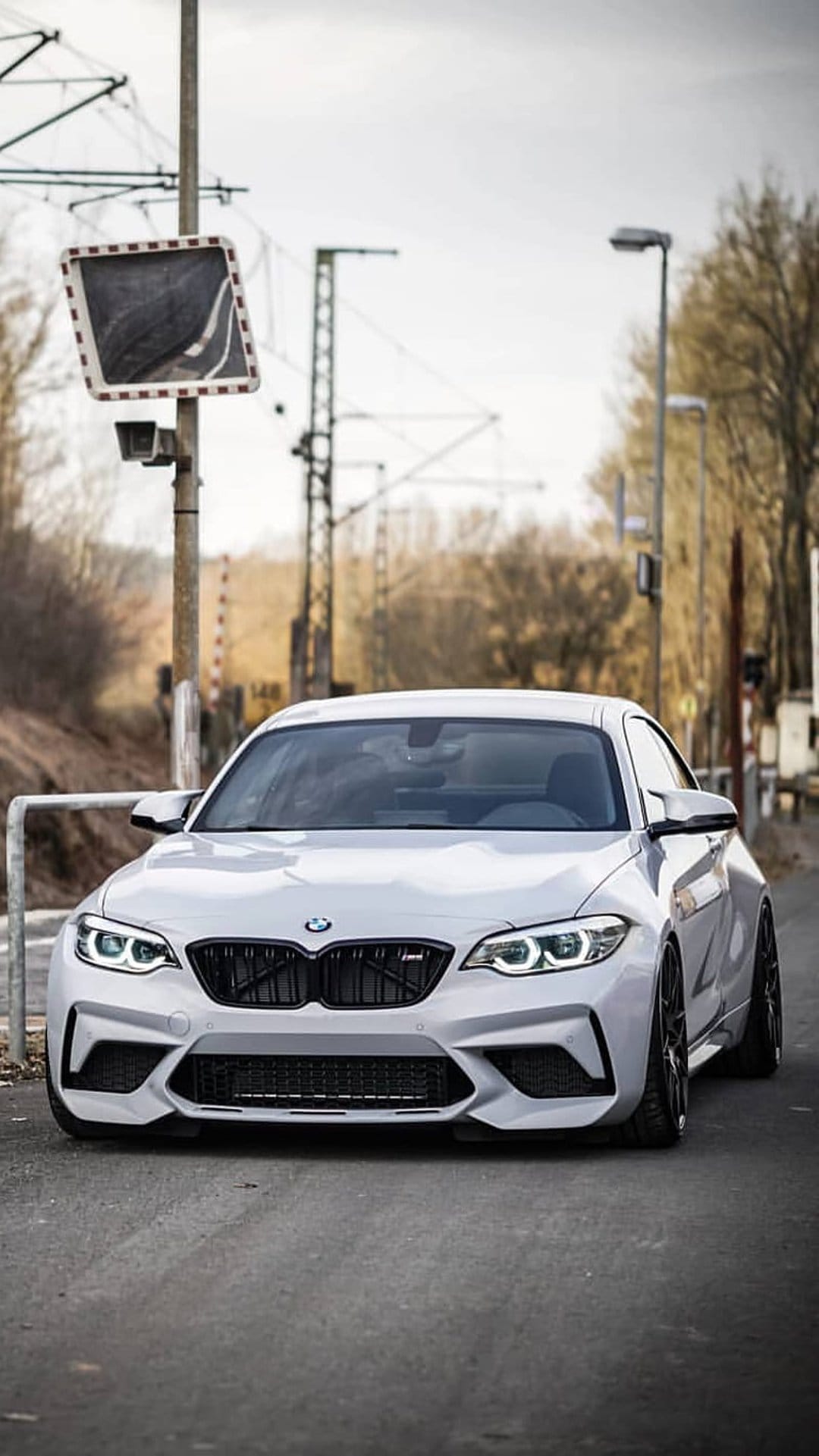 White BMW M2 sports car parked on asphalt beside railway tracks under a cloudy, overcast industrial grey sky.