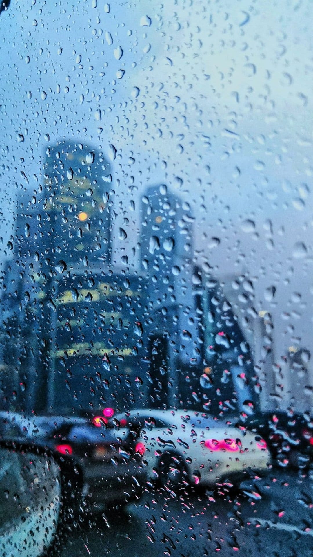 Raindrops on a windowpane blur the glowing lights of a skyscraper and evening traffic in a vertical city view.