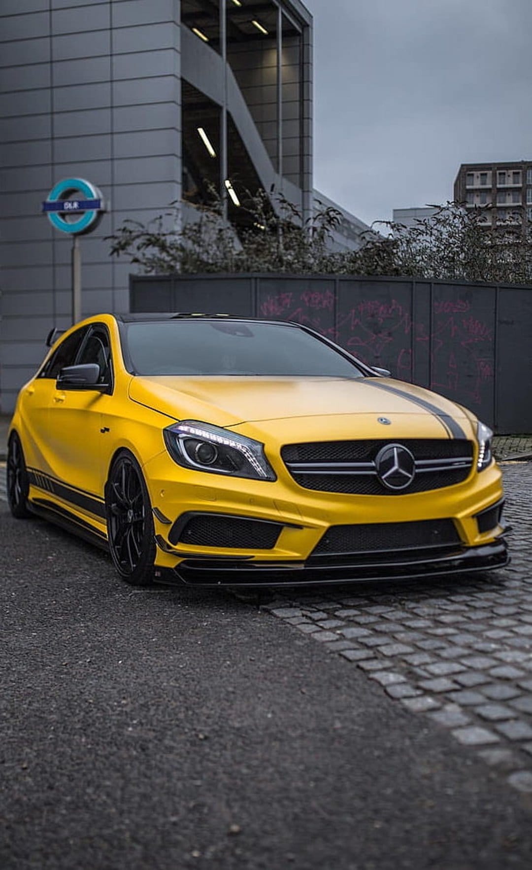 Yellow Mercedes-Benz AMG hatchback with black stripes parked on a cobblestone street near a London subway sign.