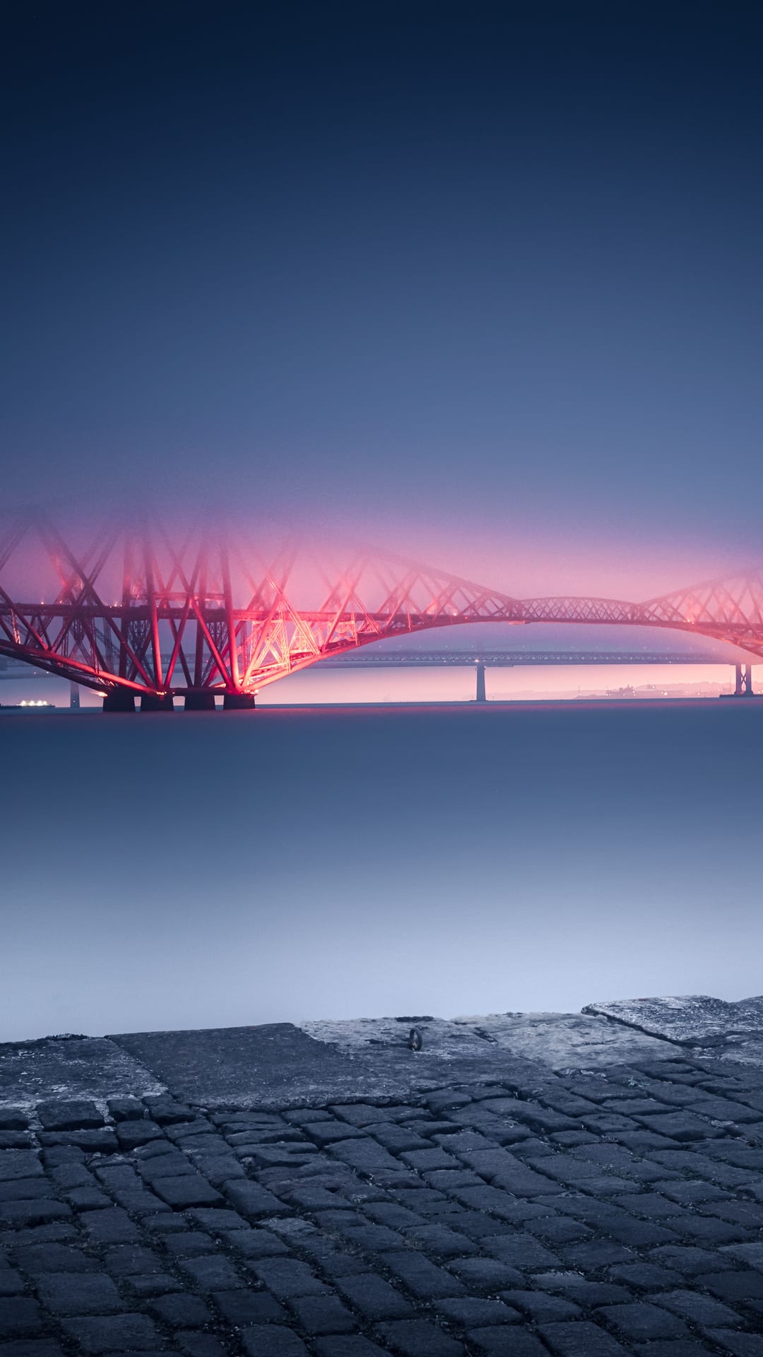 Forth Bridge glowing red under a twilight sky reflects in the dark Scottish estuary waters at dusk.