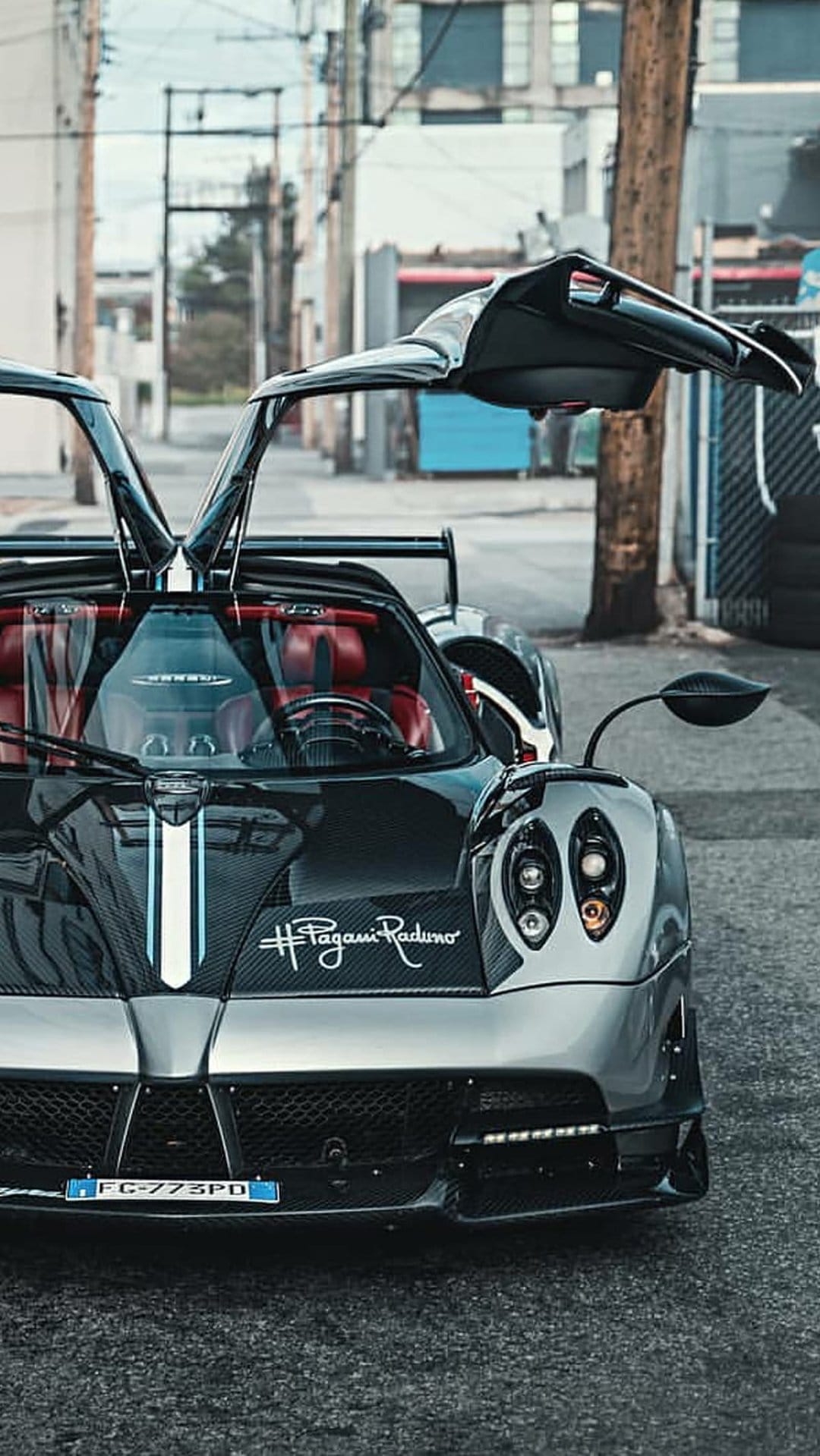 Pagani Huayra BC Roadster with open gullwing doors, red interior, and white racing stripe in a dark urban alley.