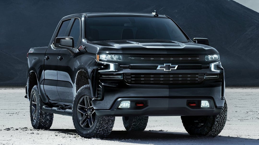 Black Chevrolet Silverado Trail Boss pickup truck with glowing headlights on a desert salt flat at twilight.