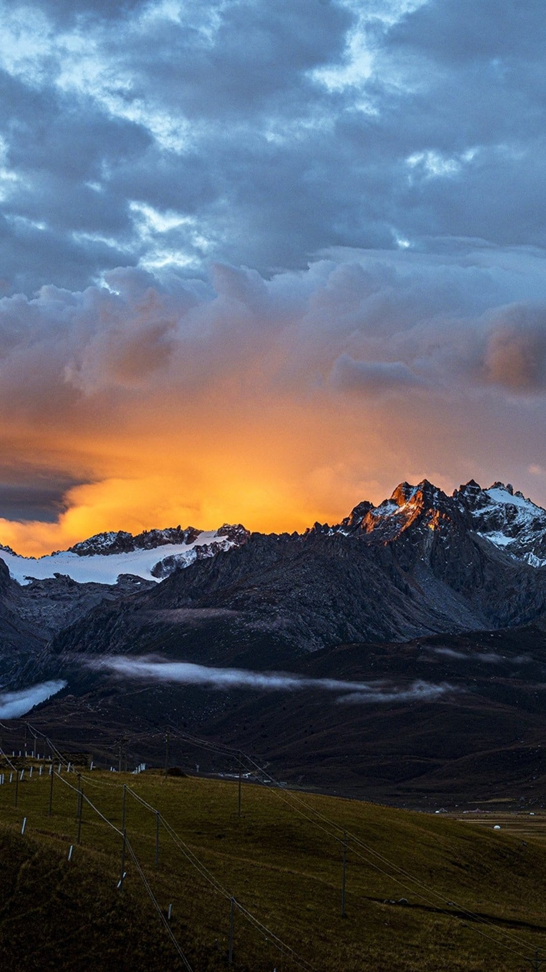 Snow-capped mountain peaks glow with orange sunset light beneath dark heavy clouds and green alpine slopes.