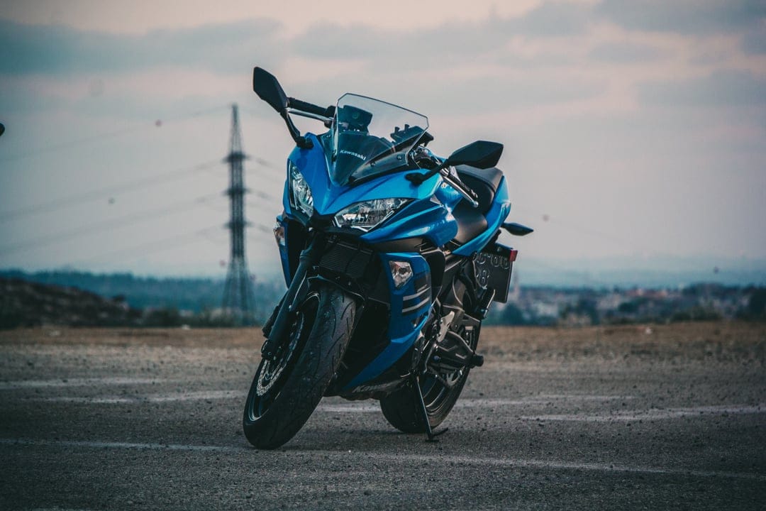 Blue Kawasaki sportbike parked on gravel with a tall industrial tower and city skyline under a cloudy sky.