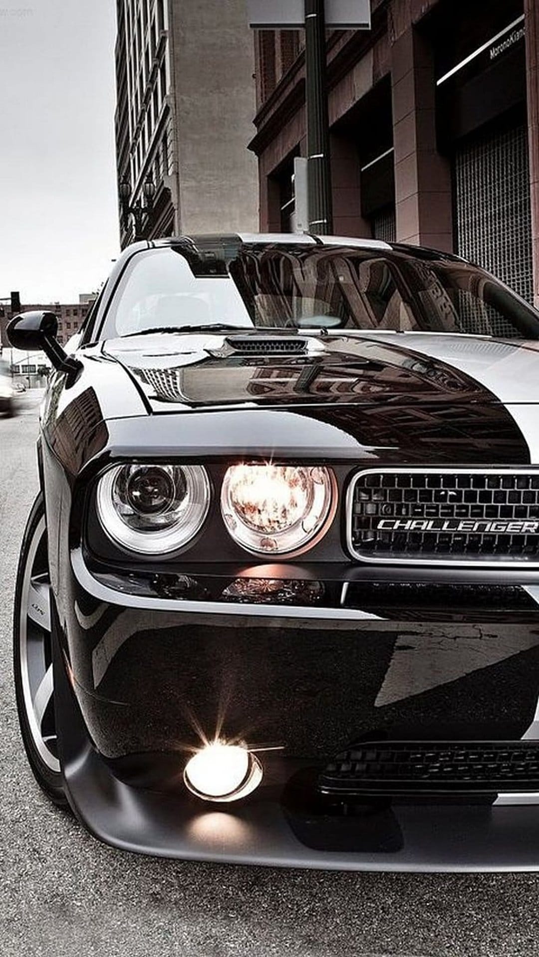 Black Dodge Challenger with a striped hood and circular headlights parked on a reflective city street at dusk.