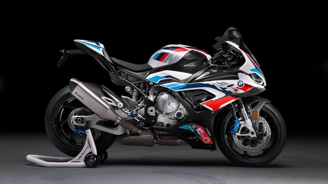 Side profile of a white and blue BMW M 1000 RR motorcycle with carbon fiber parts in a dark studio background.