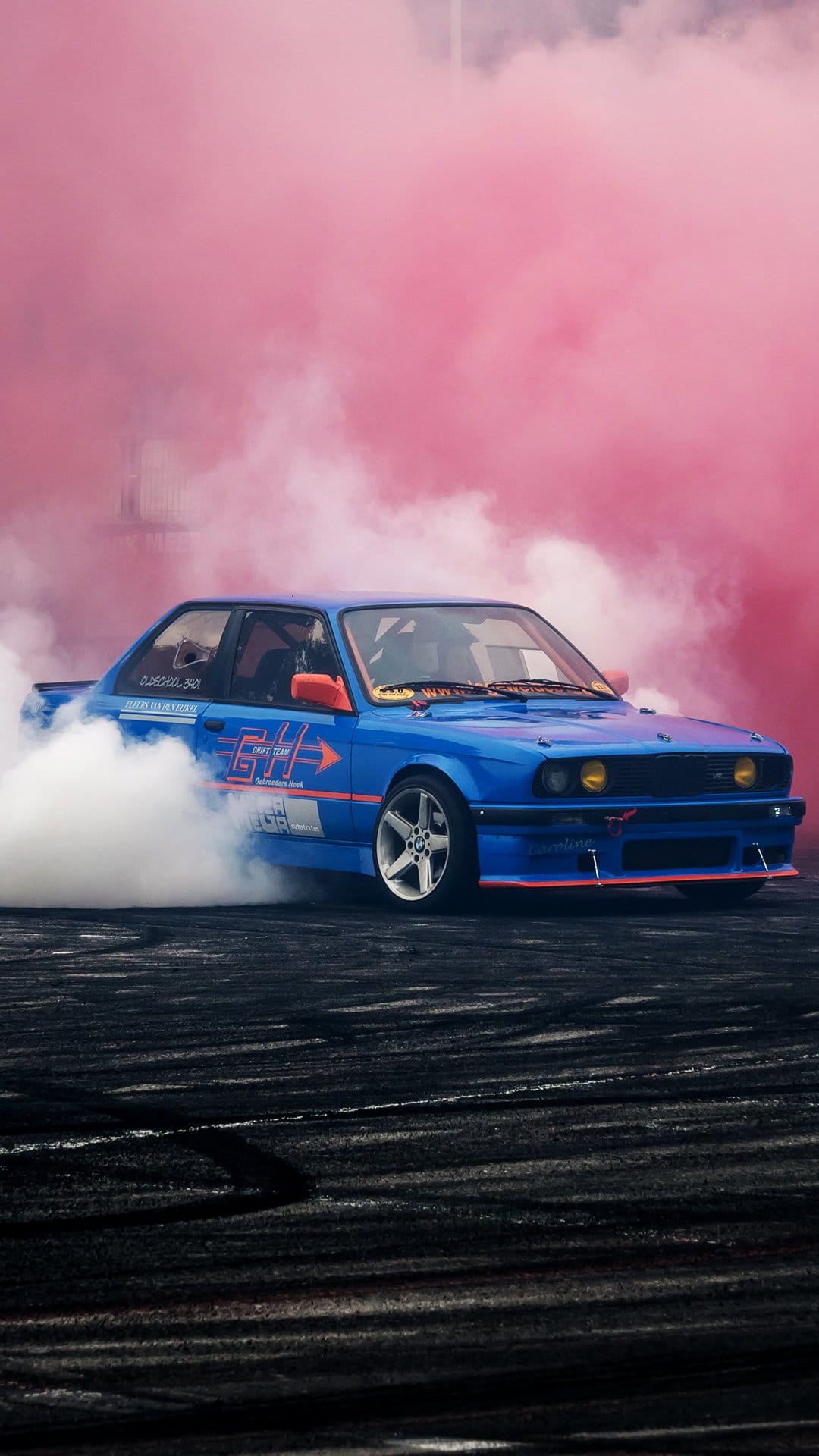 Blue drift car with orange accents skidding on dark asphalt while emitting thick white and pink tire smoke clouds.