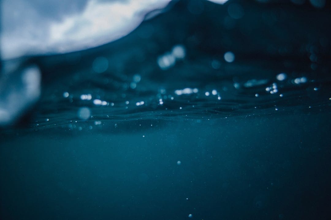 Dark blue ocean waves churn above rising air bubbles and shimmering light shafts in a deep sea environment.
