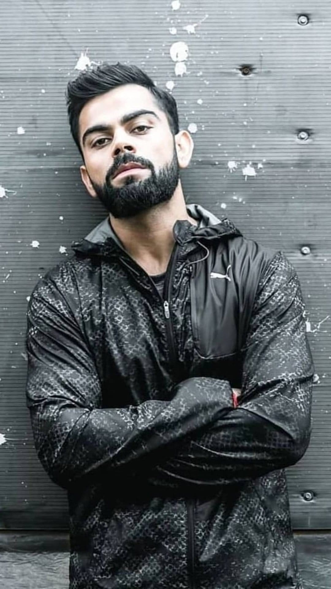 Virat Kohli in a black Puma jacket leans against a textured gray wall with his arms crossed and an intense gaze.