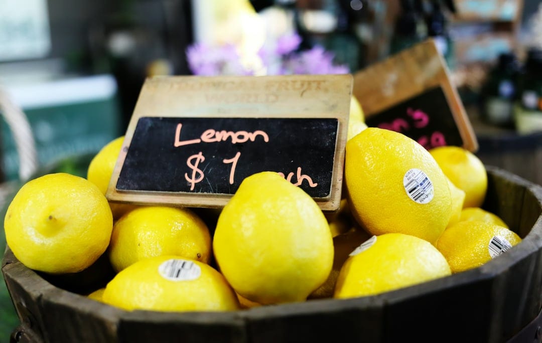 Yellow lemons in a wooden bucket next to a chalkboard sign at a market with a soft blurred background.
