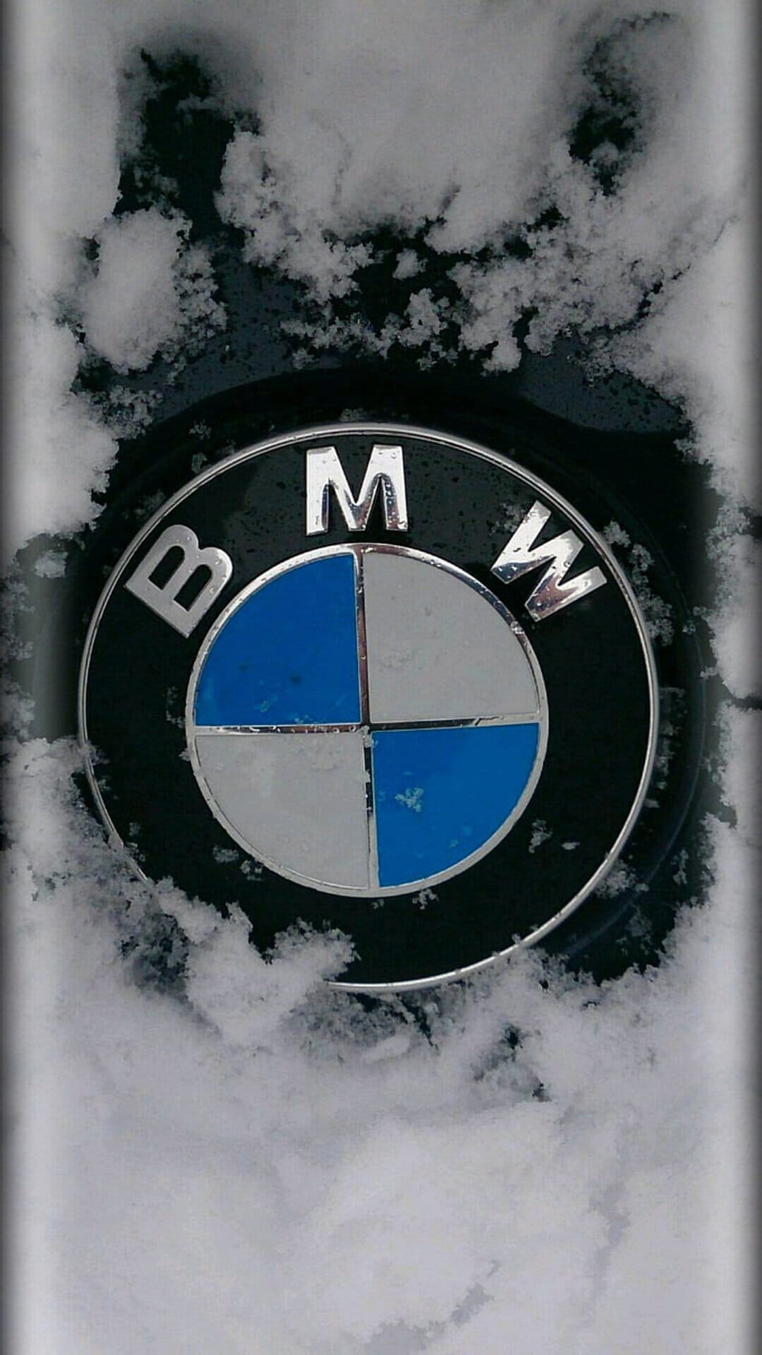 BMW emblem partially covered in white snow crystals against a dark textured car grille in frozen winter weather.