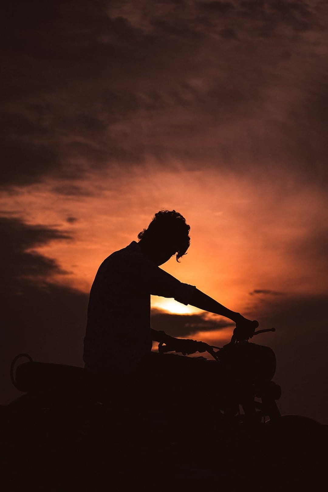 Biker silhouette with curly hair on a motorcycle against a dark orange sky with textured sunset clouds.