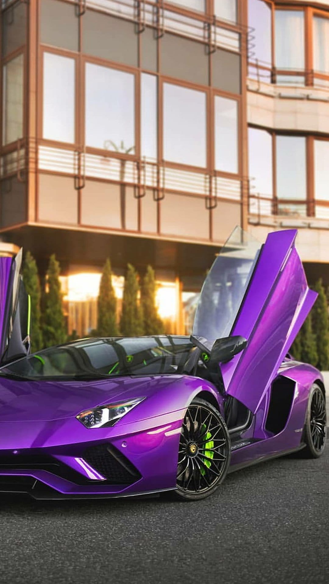 Purple Lamborghini Aventador with raised scissor doors, green calipers, and modern architecture at sunset.