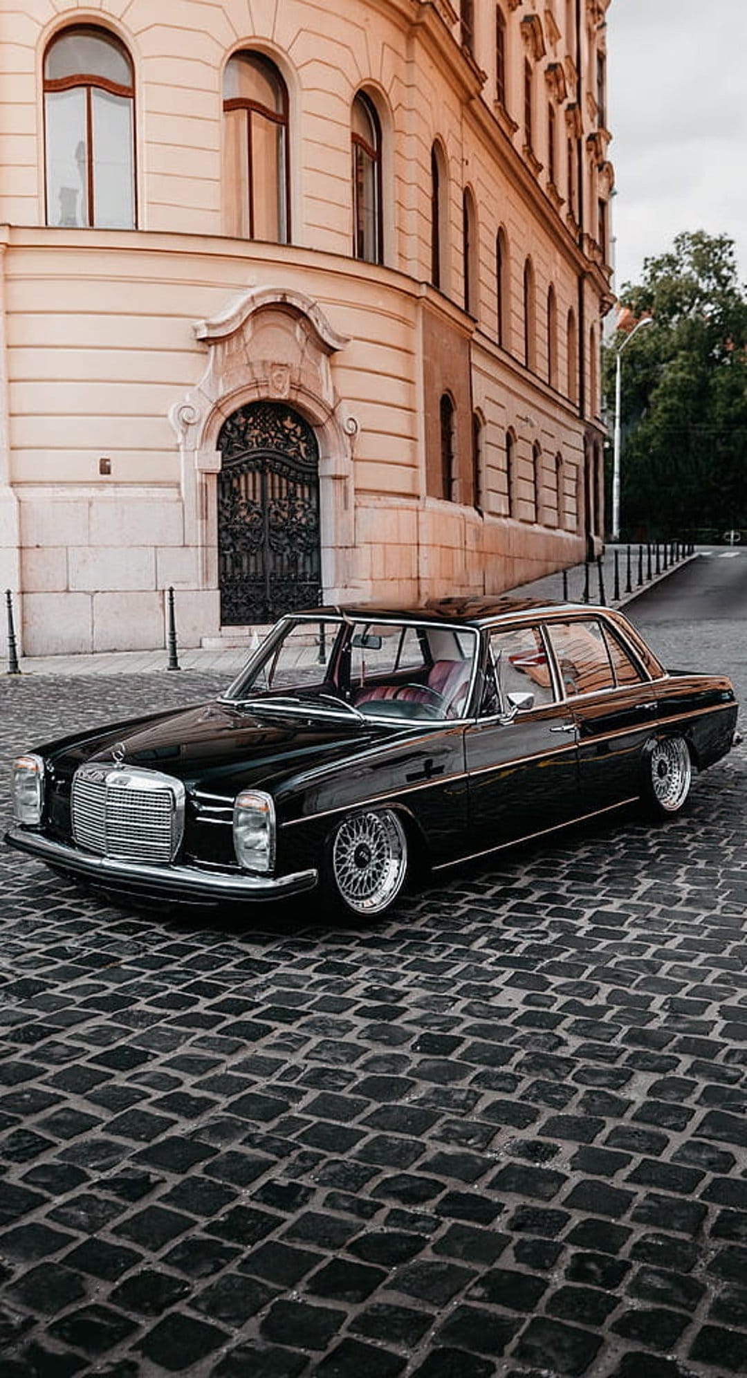 Black vintage Mercedes-Benz W116 sedan with chrome trim parked on a textured European cobblestone street.