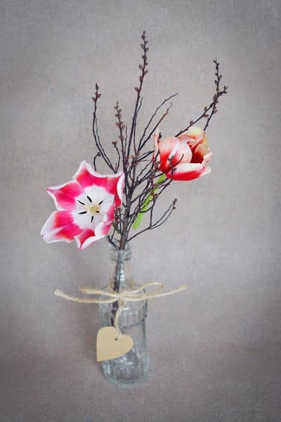 Pink and white tulips in a glass jar with a wooden heart tag and twigs against a textured beige background.