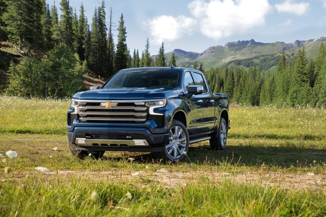 Blue Chevrolet Silverado High Country parked in a green mountain meadow with pine trees and a clear sky.