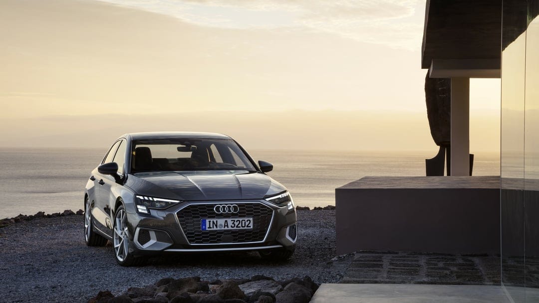 Charcoal Audi A3 sedan on a gravel coast at sunset with a golden horizon and a modern building fragment.