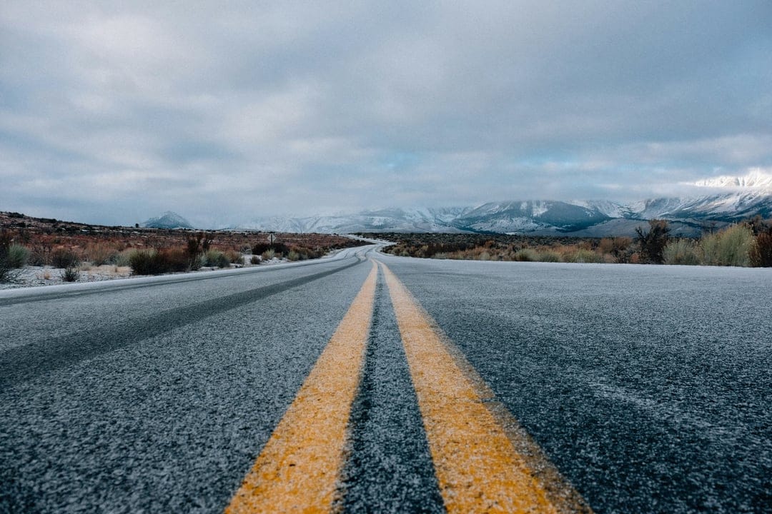 Asphalt road with double yellow lines leads toward jagged snow-covered mountains under a heavy cloudy gray sky.