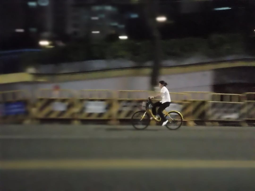 Cyclist in a white top rides a yellow bike past streetlights with motion blur on a dark city road at night.