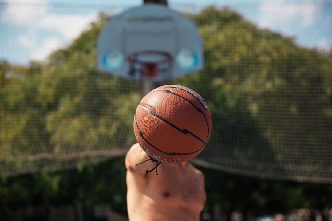 Textured basketball spinning on a single finger with a blurred backboard and trees in a sunny outdoor setting.