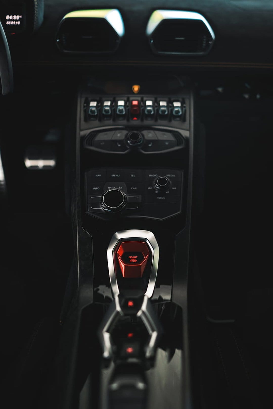 Red engine start button with a flip cover sits amidst the carbon fiber console and vents of a supercar cockpit.