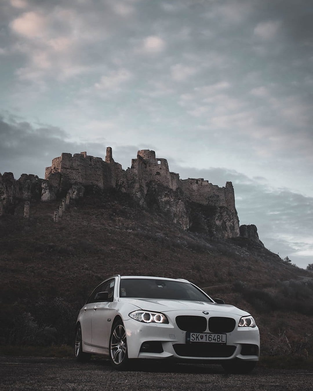 White BMW station wagon parked before crumbling stone castle ruins under a dramatic, moody overcast sky.