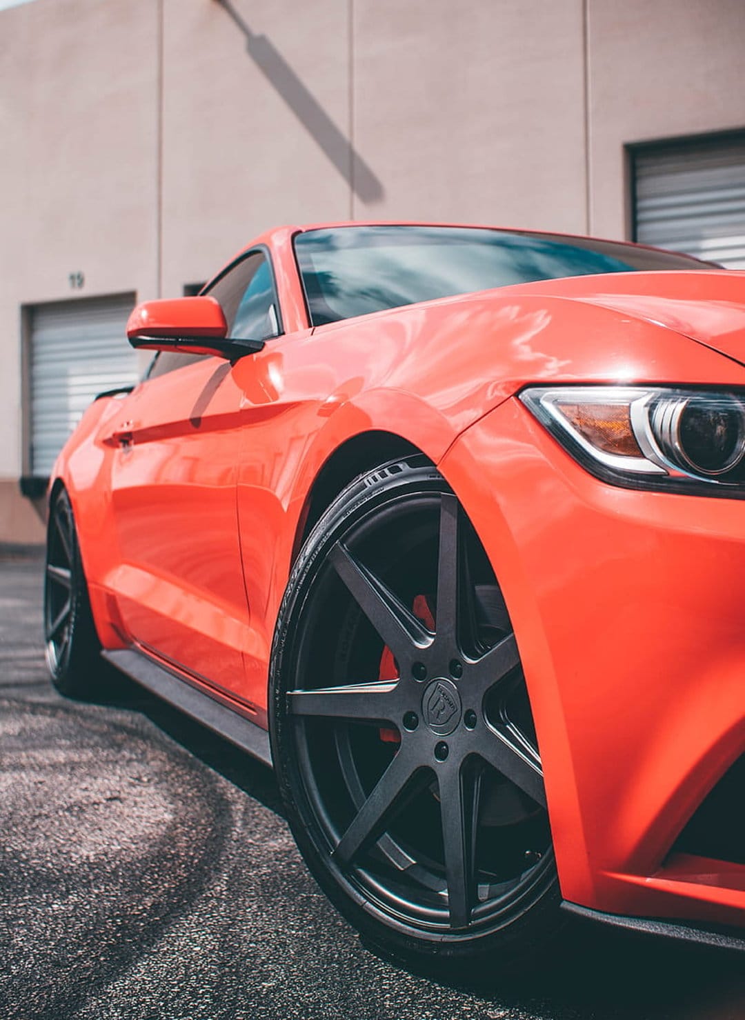 Orange Ford Mustang with custom black wheels and red brake calipers parked in front of a modern urban building.