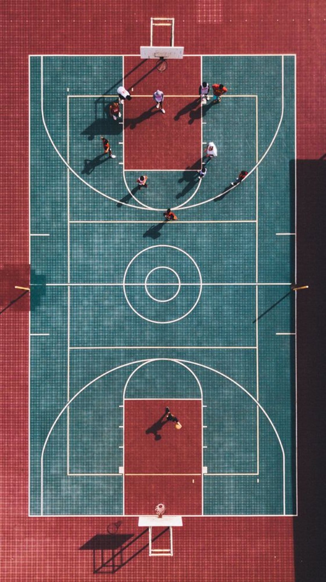 Athletes play basketball on a red and teal outdoor court viewed from a direct top-down aerial perspective.