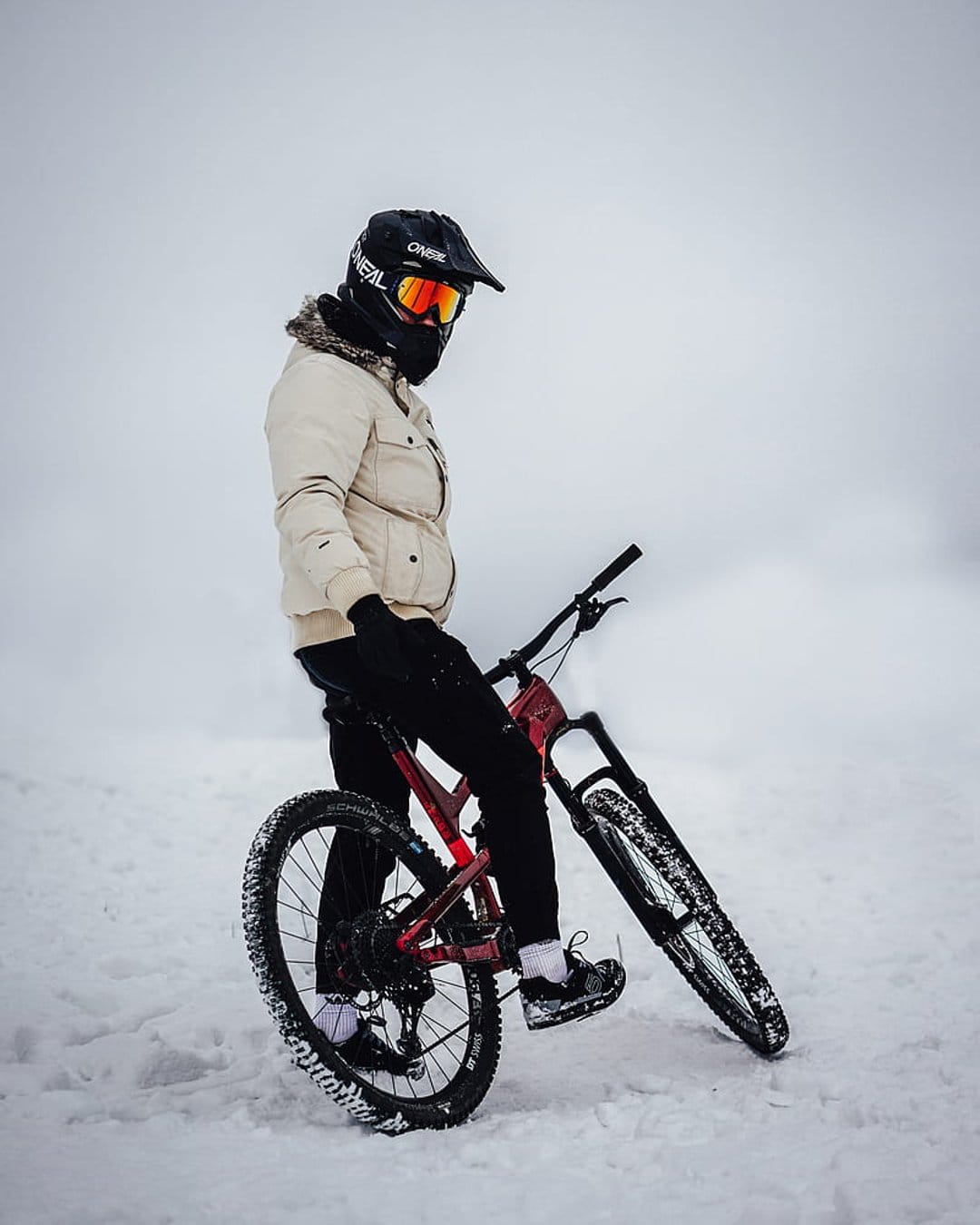 Mountain biker in a full-face helmet with a red bike stands on a snow-covered trail during a winter ride.