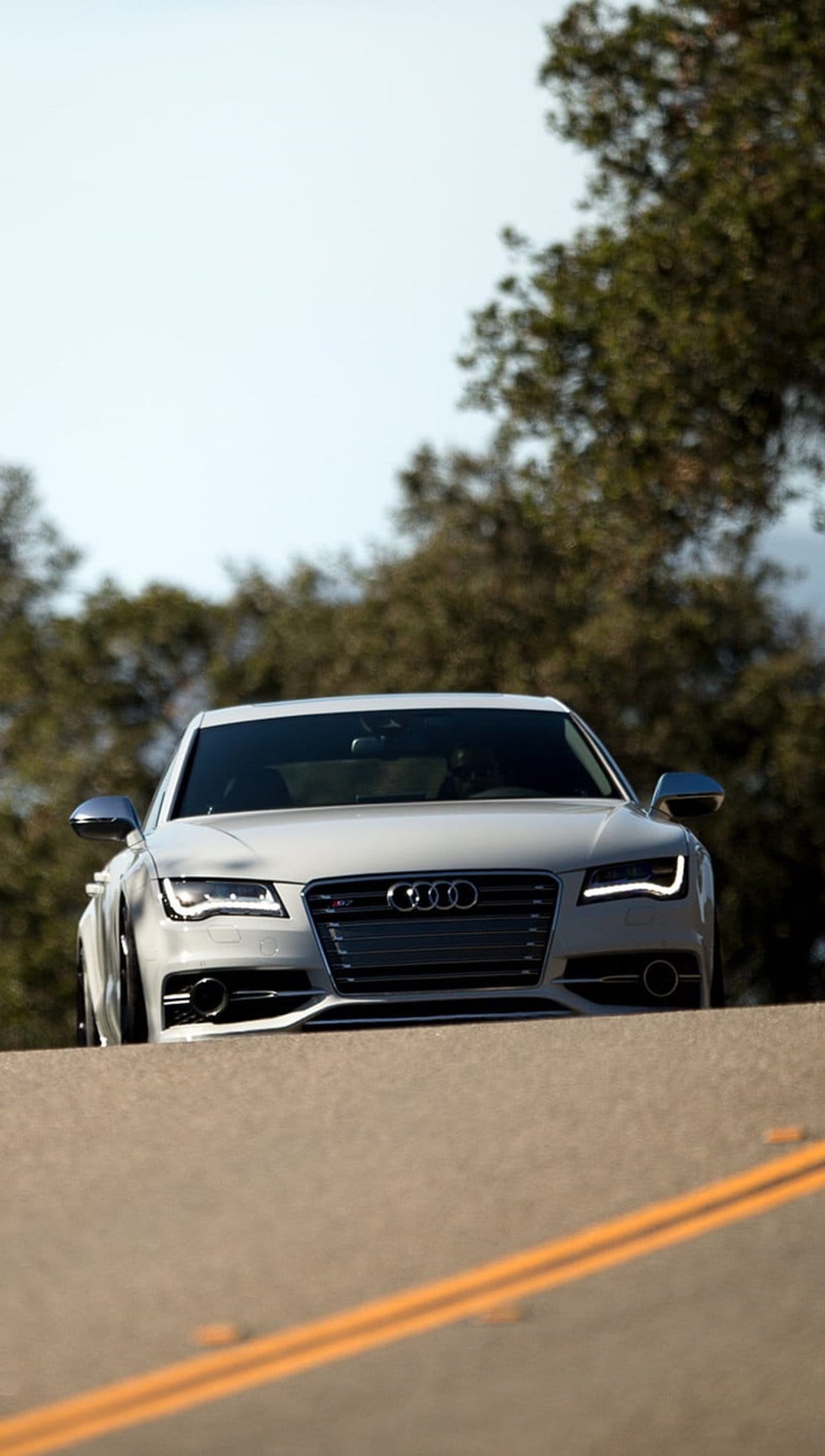 White Audi S7 sedan driving on a scenic asphalt road with yellow markings and trees in the background.