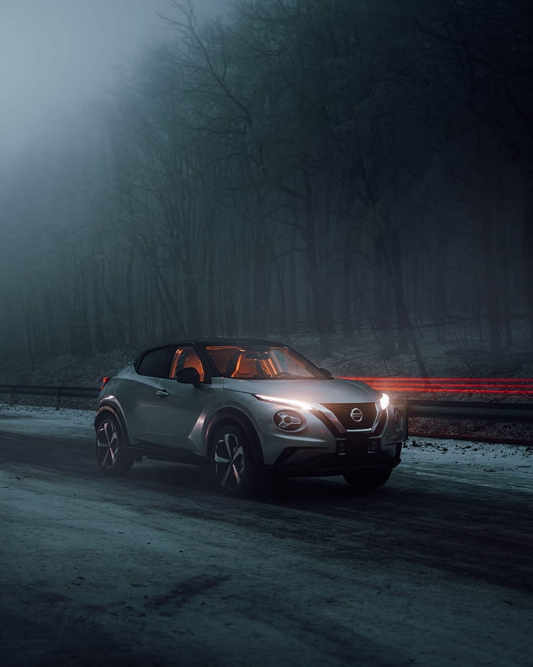 Nissan Juke with glowing headlights traverses a snowy forest road under dense fog and dark night conditions.