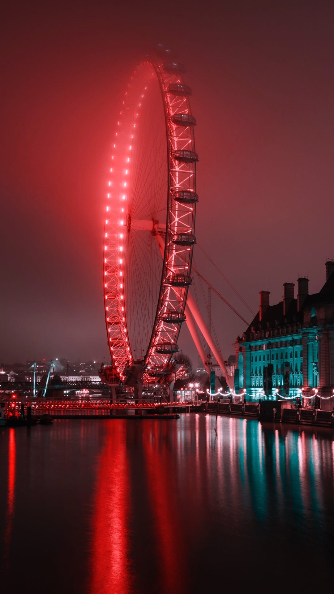 London Eye glowing bright red through thick fog with reflections on dark river water and distant teal lights.