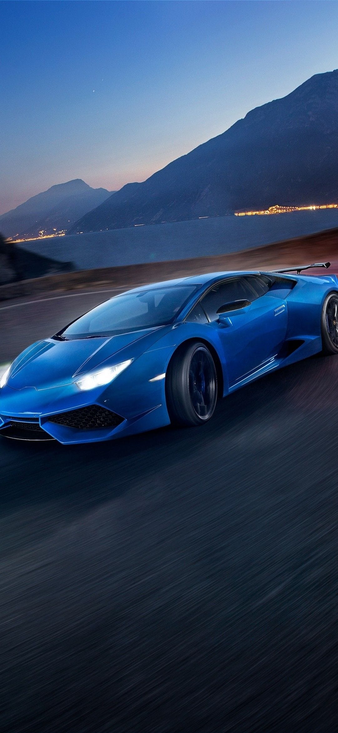 Blue Lamborghini Huracán speeding on a coastal mountain road at dusk with glowing headlights and city lights.