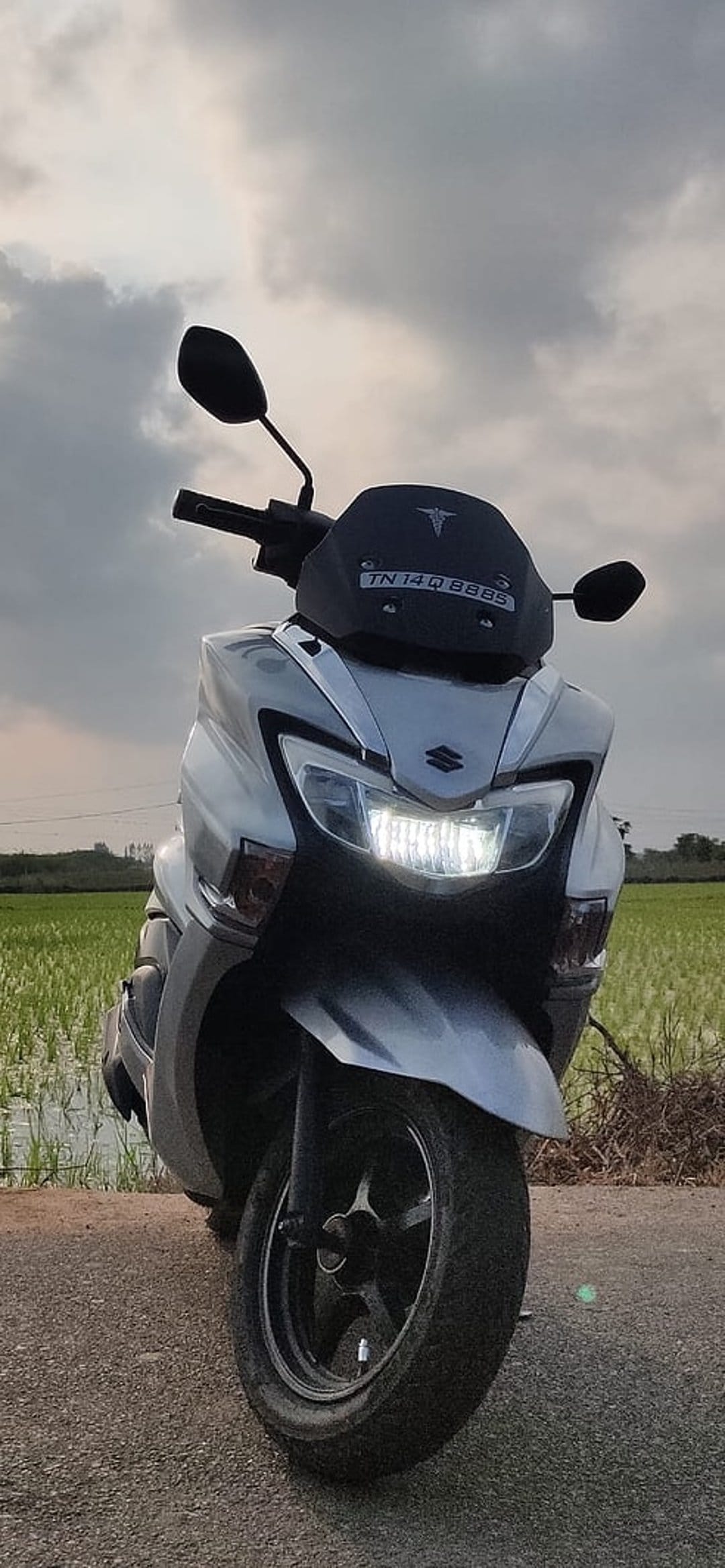 Suzuki Burgman scooter with glowing headlights parked beside a lush green paddy field under a cloudy sunset sky.
