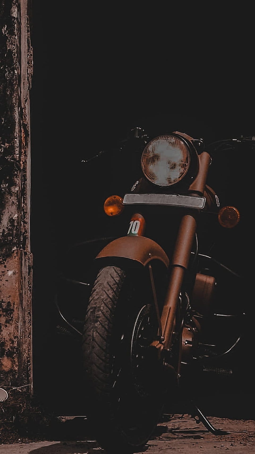Vintage motorcycle headlight and orange turn signal emerge from dark shadows next to a textured wooden post.