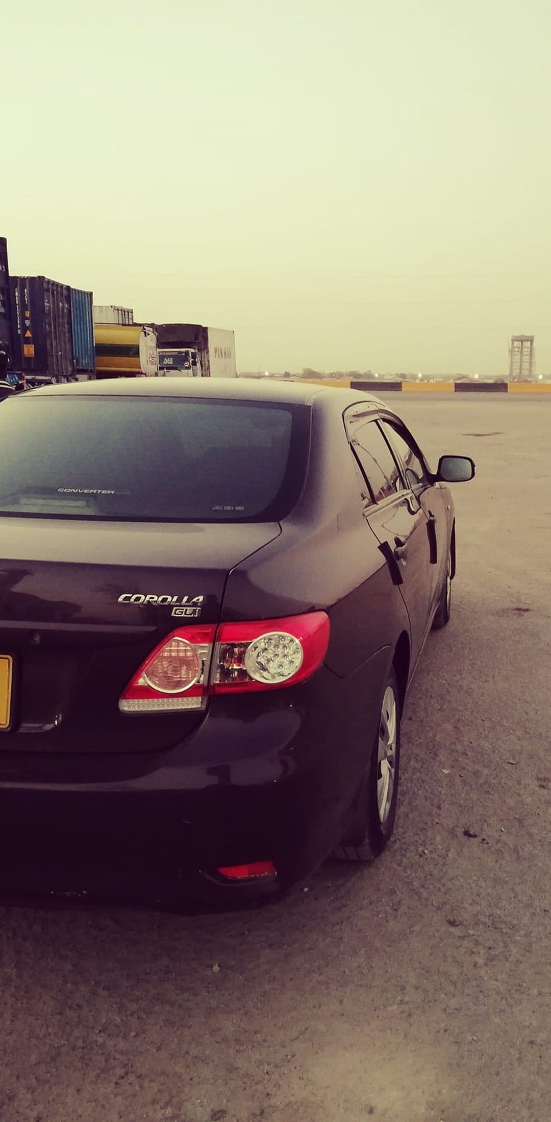Dark Toyota Corolla GLi rear view in a shipping yard with stacked containers under an overcast, hazy sky.