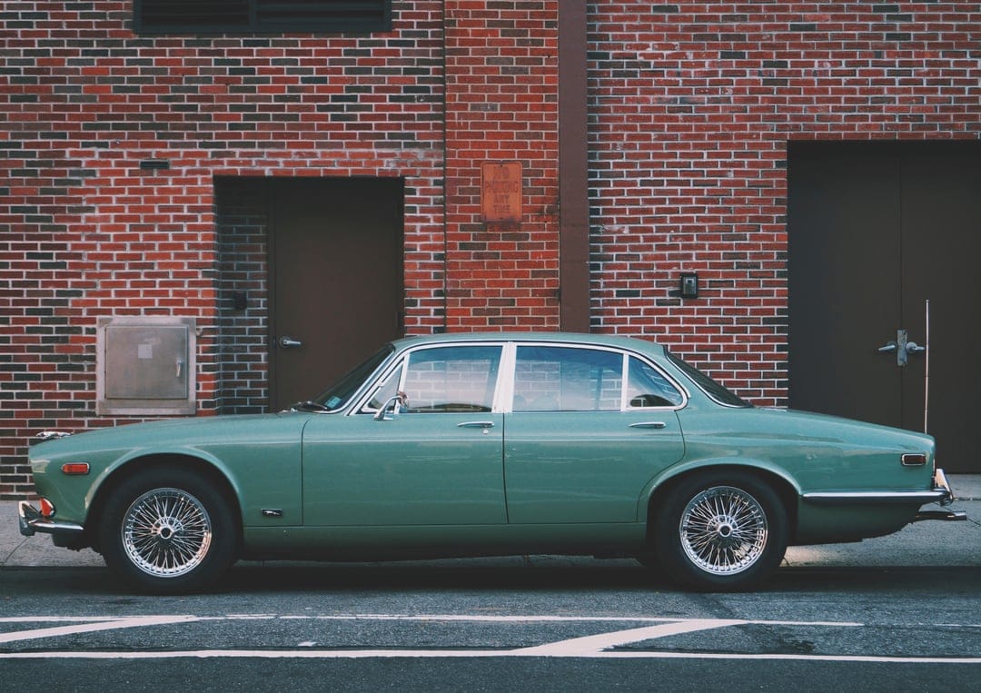 Vintage green Jaguar sedan with chrome accents parked next to a weathered red brick building with brown doors.