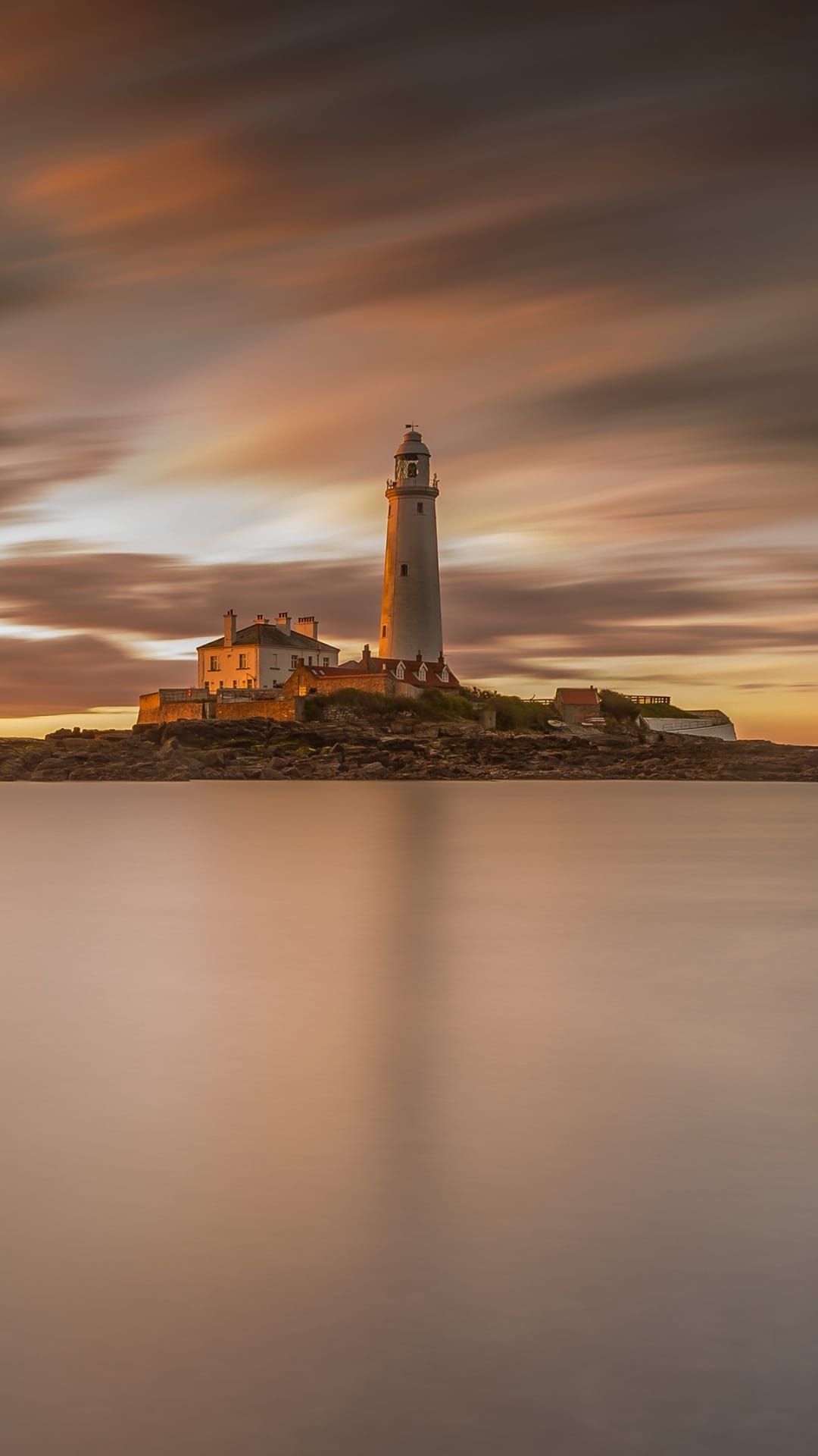 St. Mary's Lighthouse stands on rocks under an orange sunset sky with long-exposure blurred clouds and water.