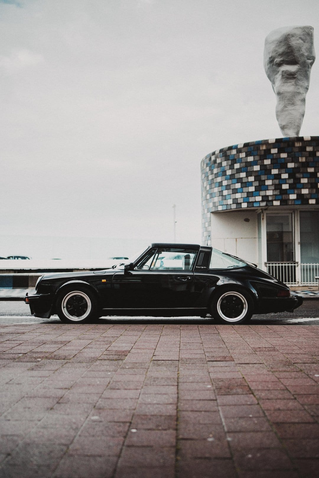 Black Porsche Targa parked on a cobblestone street with a blue mosaic building and abstract sculpture behind.
