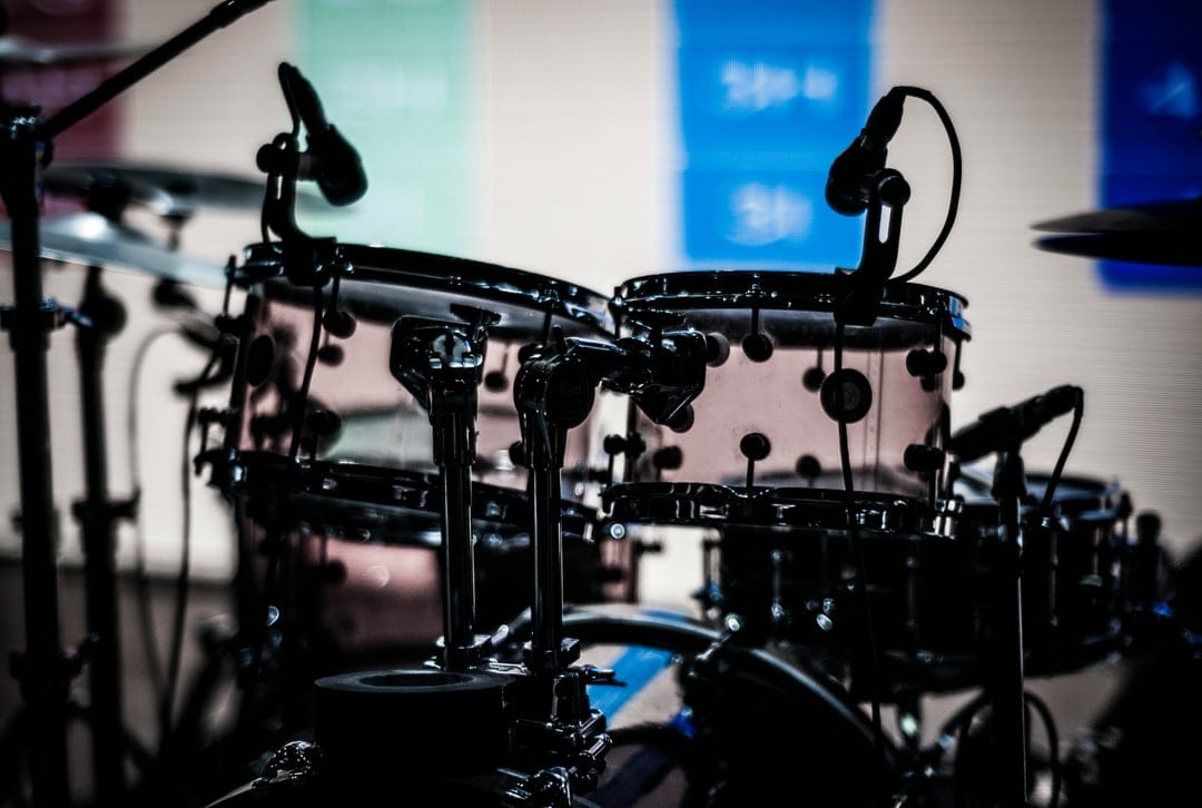 Transparent drum kit with microphones and chrome hardware under dim stage lights with a colorful blurred background.
